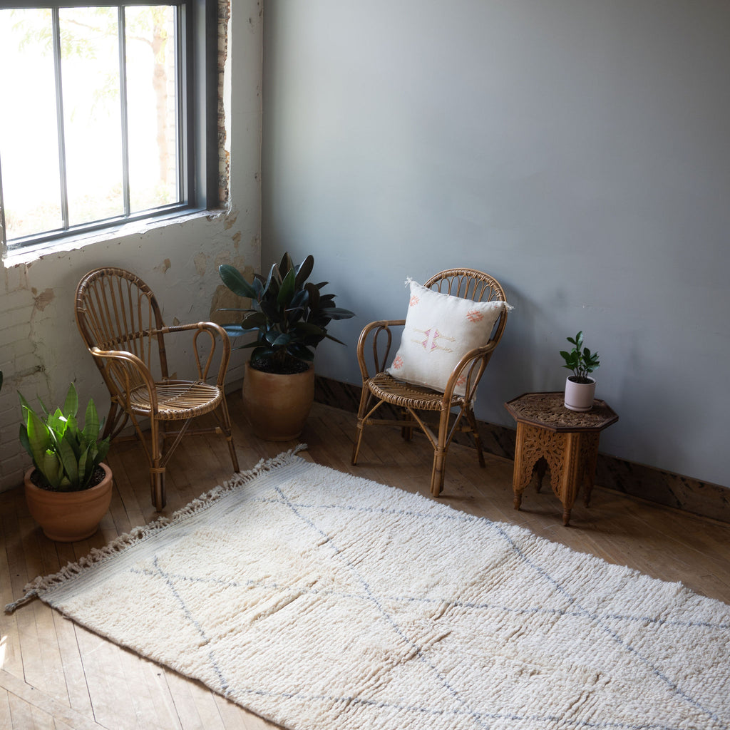 A diagonal view of a high pile Moroccan Beni Ourain rug with classic cream and gray diamond design and tasseled/fringed edge surrounded by two rattan chairs, a side table, and a potted plant. Wood floors.