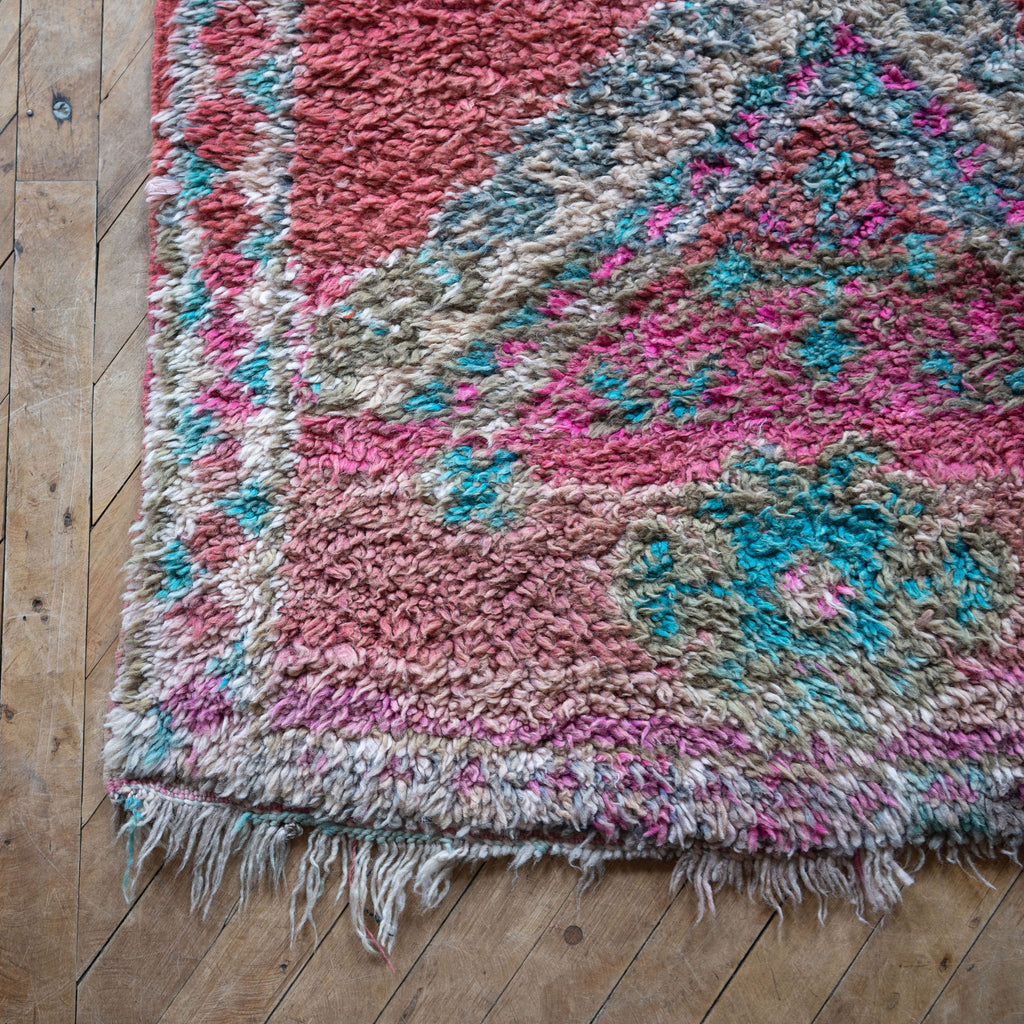 Close up of the worn fringed corner of a vintage Moroccan Benimguild rug with a bold diamond pattern in coral, turquoise, cream and pink. Wood floor.