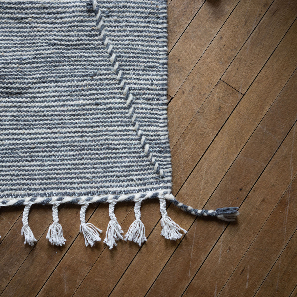 Close up of the tasseled corner of a flat woven wool Moroccan Zanafi rug with a subtle stripe design in slate gray and white with thicker cords woven through in a large triangle design. Wood floor.
