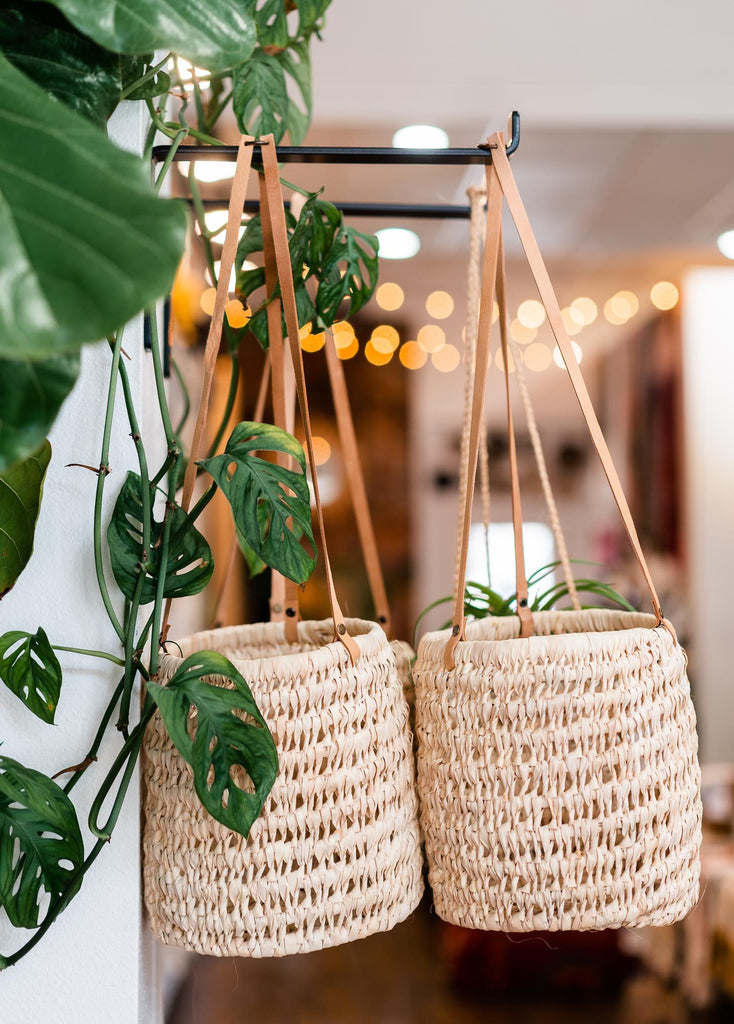 Two palm fiber loosely woven straight sided flat bottomed baskets with brown leather hanging straps. Hanging from a black iron shelf bracket in front of a Monstera Adansonii.