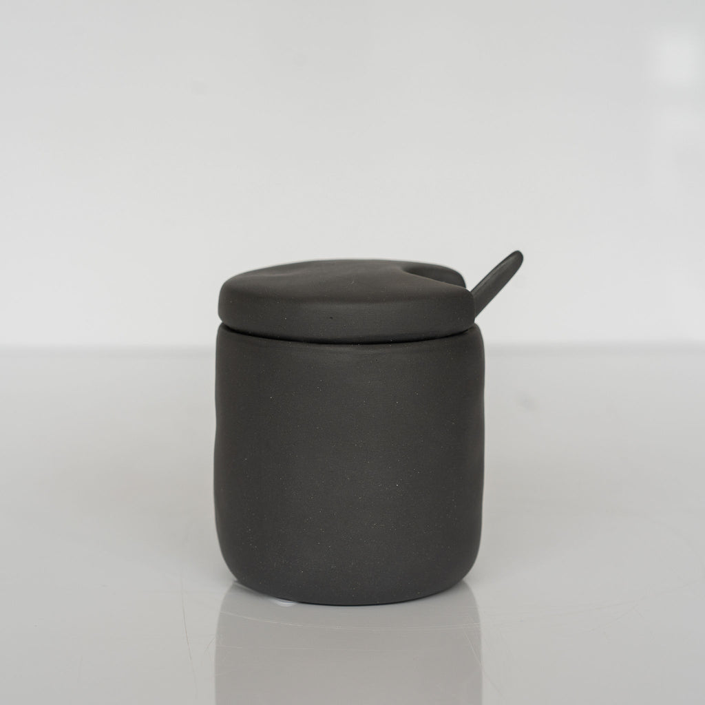 Small dark gray stoneware cellar with notched lid and spoon sits on a white background.