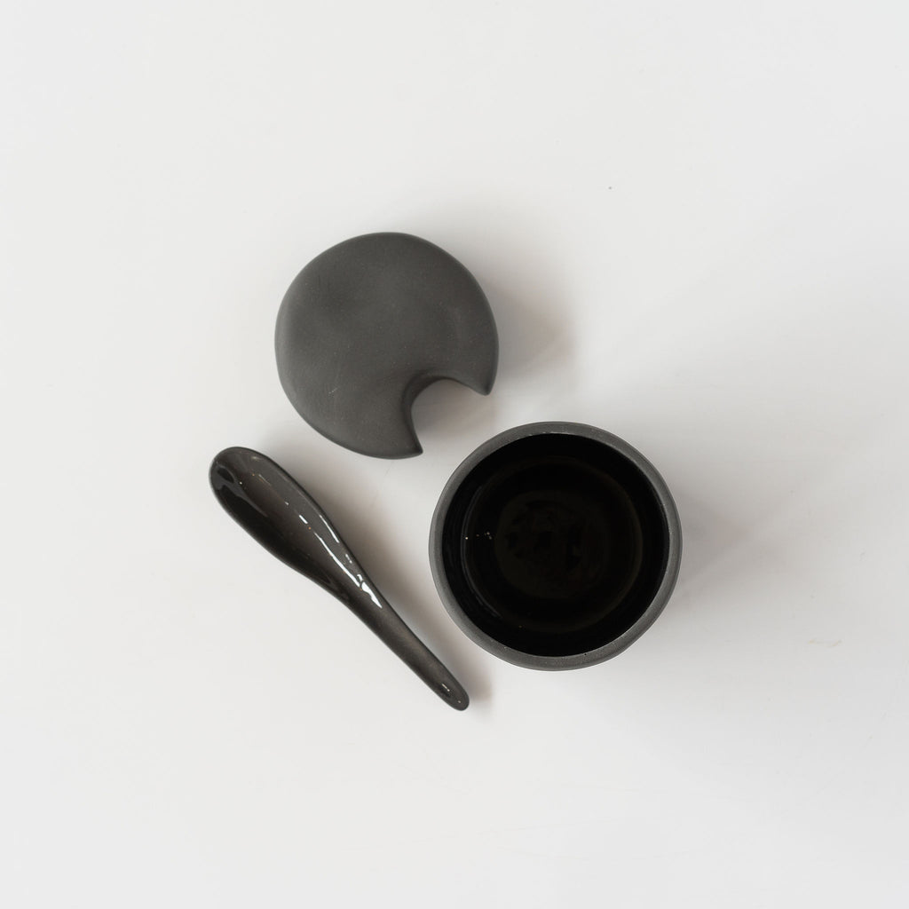 Top view of small dark gray stoneware cellar with notched lid and spoon sits on a white background.