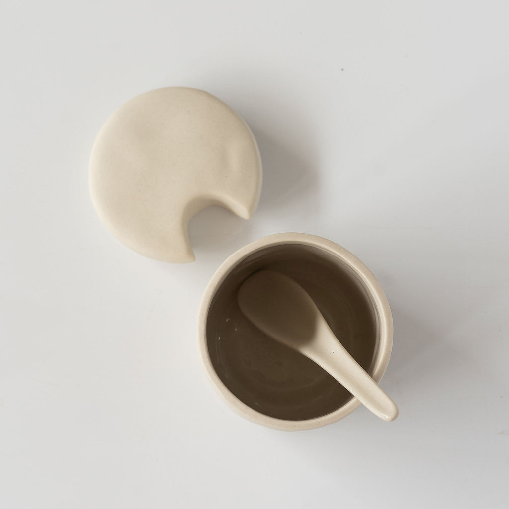 Top view of small cream colored stoneware cellar with notched lid and spoon sits on a white background.
