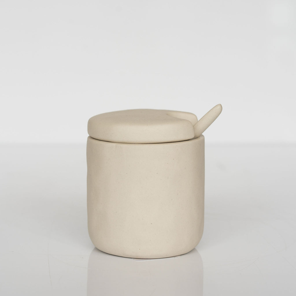 Small cream colored stoneware cellar with notched lid and spoon sits on a white background.