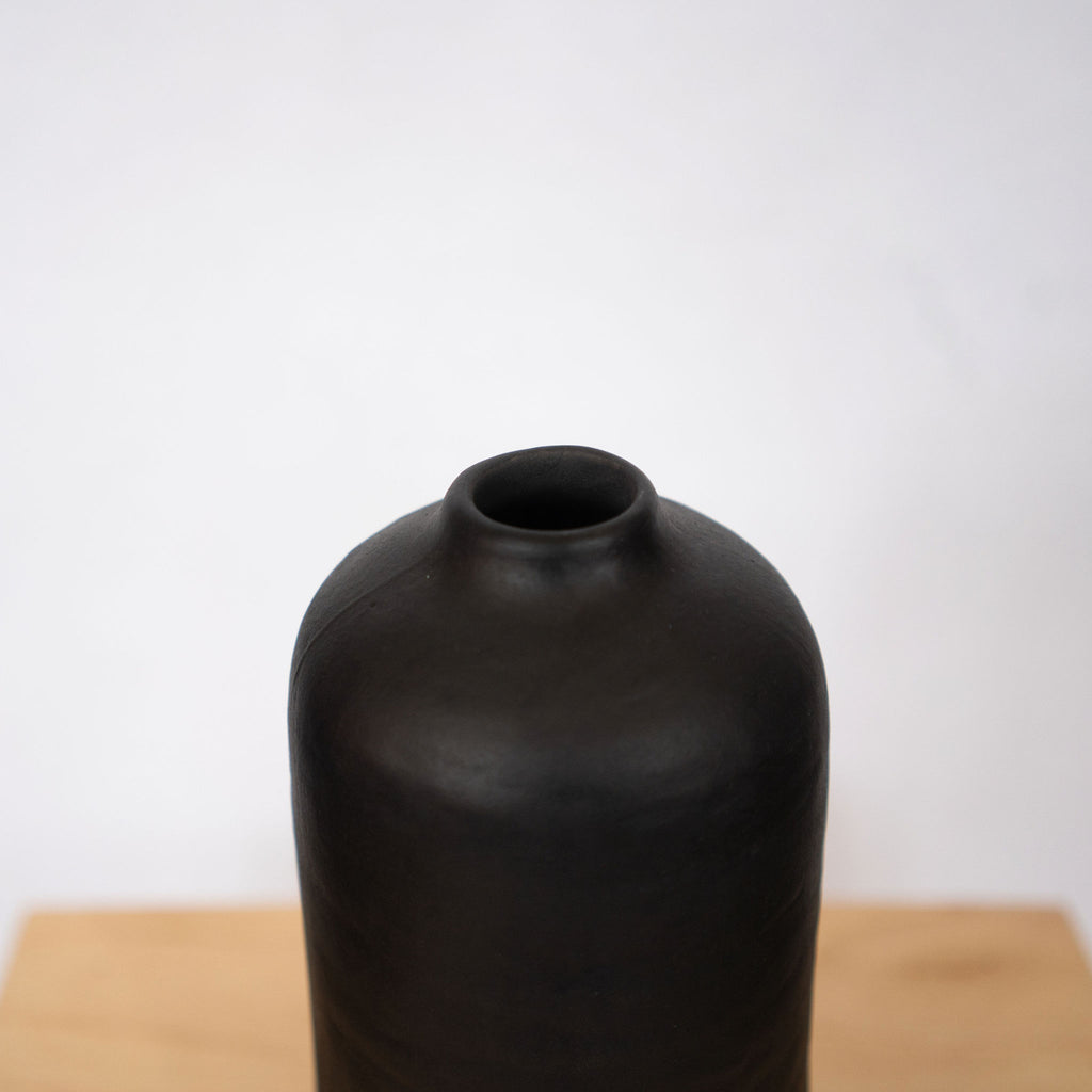 A tall straight sided black ceramic vase with small neck sits on top of a wood platform in front of a light gray background.