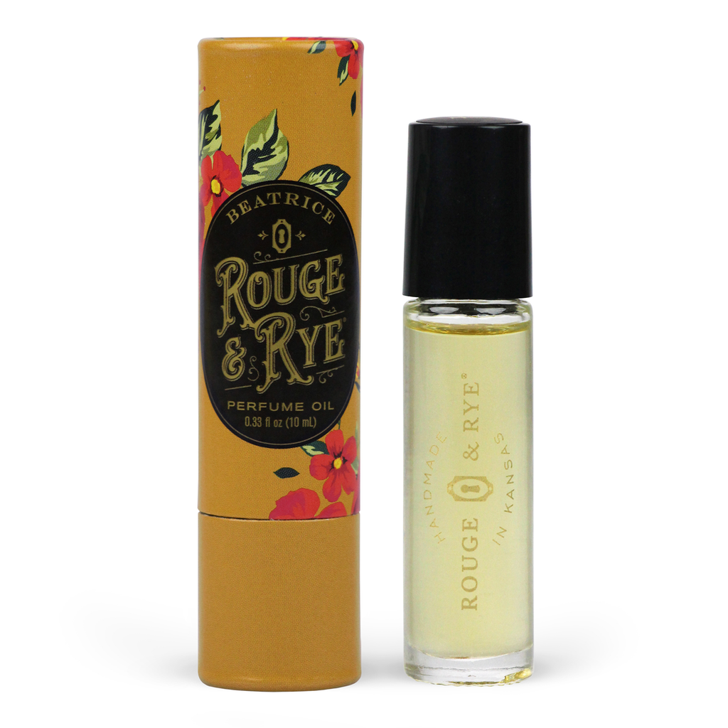 Golden Beatrice perfume oil roller in clear glass bottle with black plastic cap next to mustard floral packaging. White background.