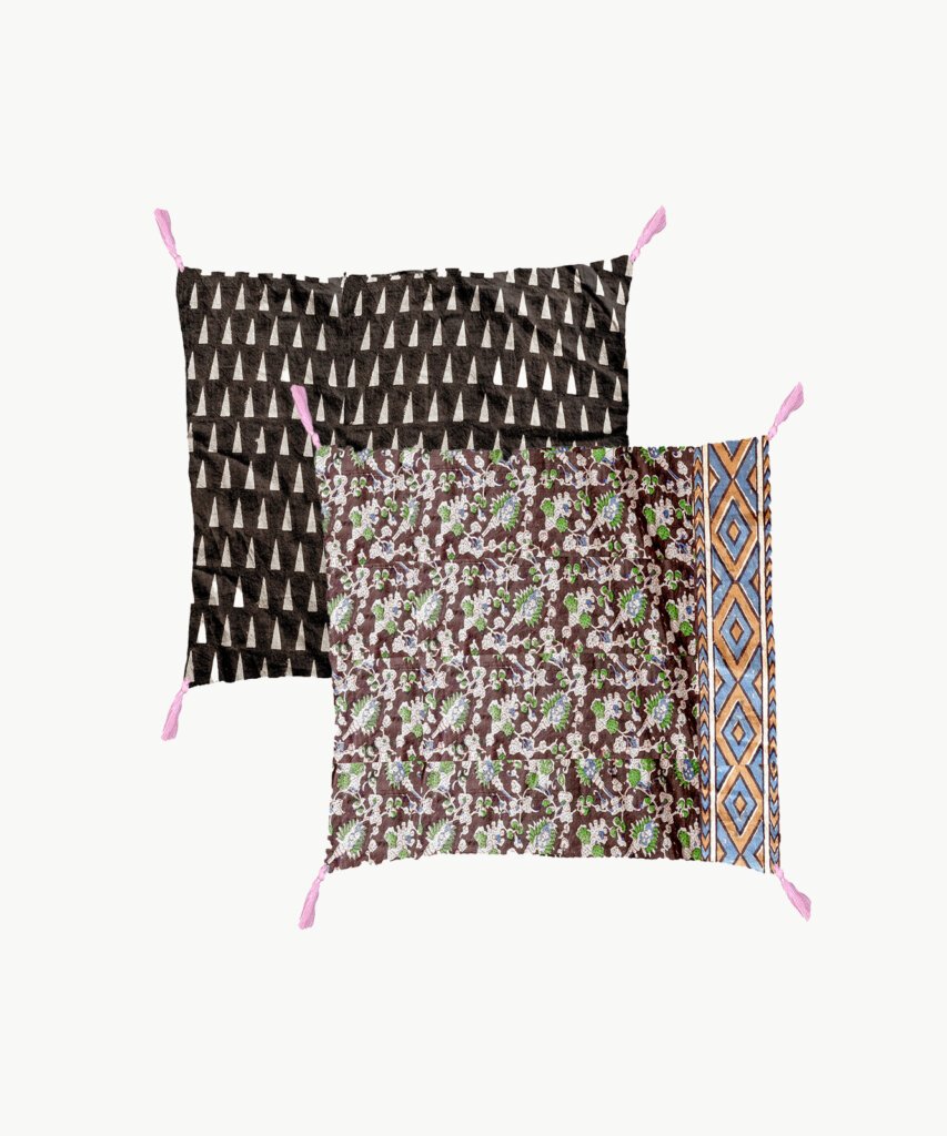 Two sides of reversible pillow showing. Four purple tassels at each corner. One side is brown background with cream triangle print. Other side is floral print with one geometric edge.
