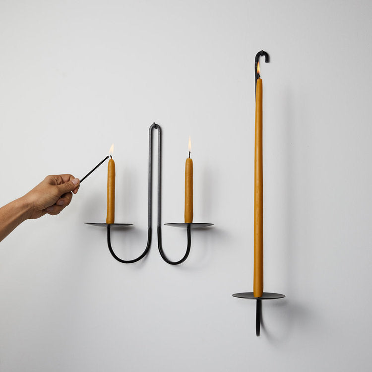 Two black iron taper candle sconces hang on a white wall holding tan taper candles. A hand is lighting one candle. 