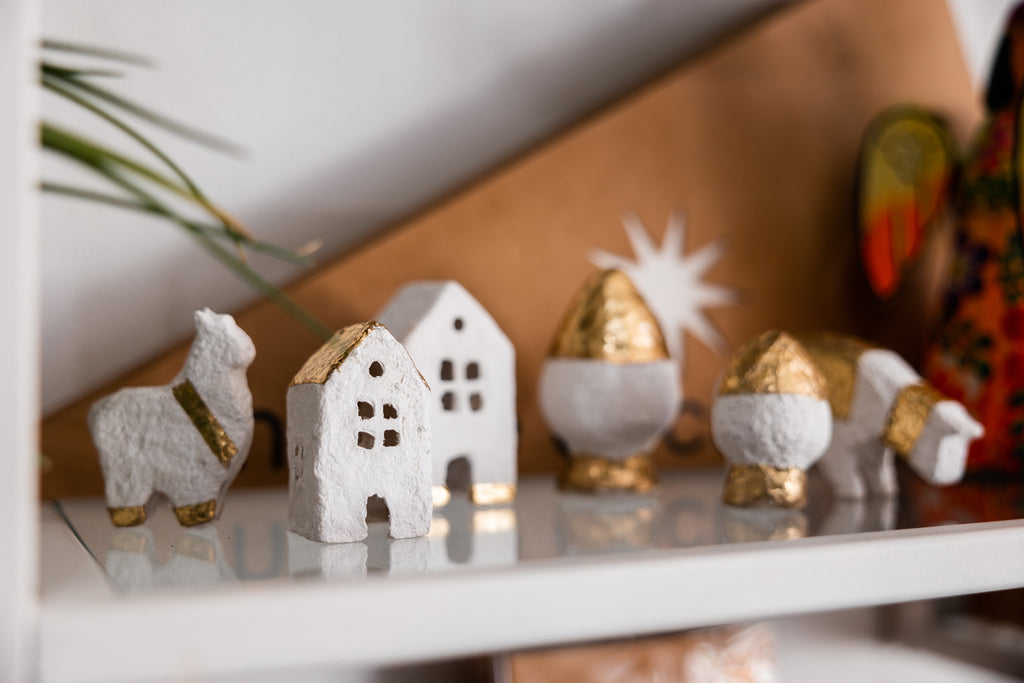 White and gold leaf cotton mache Christmas village with two houses, two trees, one cow and one llama. Sits on a glass and white shelf.