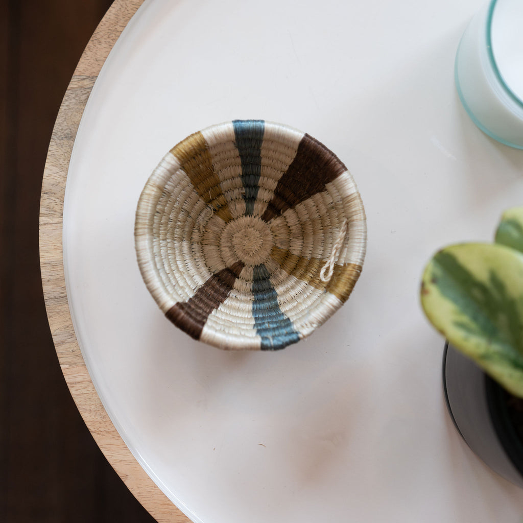 Handwoven Rwandan sweetgrass small trinket bowl in stripes in tans, cream, brown, and blue. Sits on a mango wood and white enamel tray in front of a candle and potted plant.