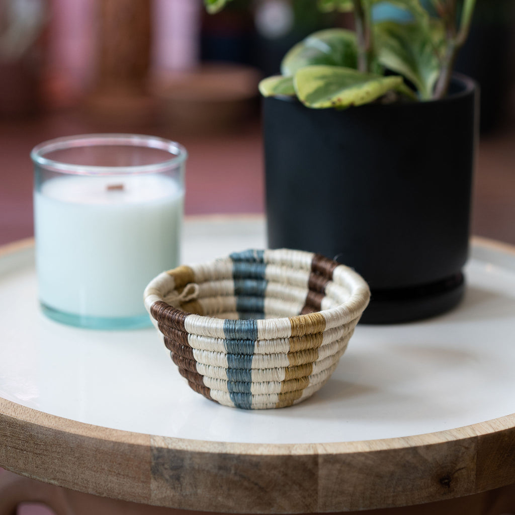 Handwoven Rwandan sweetgrass small trinket bowl in stripes in tans, cream, brown, and blue. Sits on a mango wood and white enamel tray in front of a candle and potted plant.